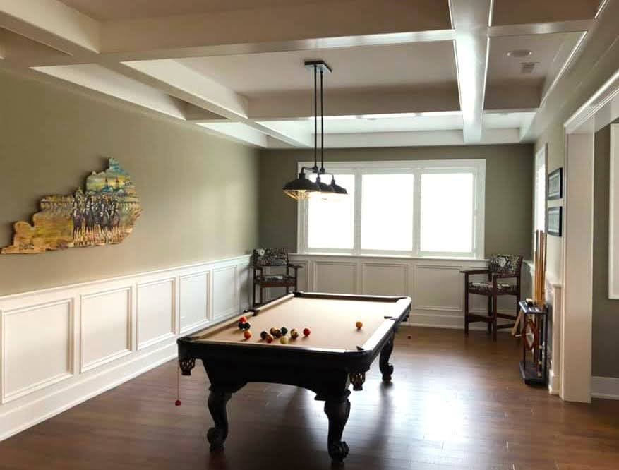 A sunlit room with a pool table, balls scattered on felt, wood floors, and wall art of a ship on the left wall.