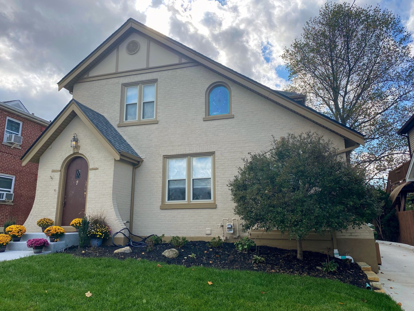 A two-story beige house with a freshly painted brown door and small windows, surrounded by a well-manicured lawn with yellow flowers and a small tree.