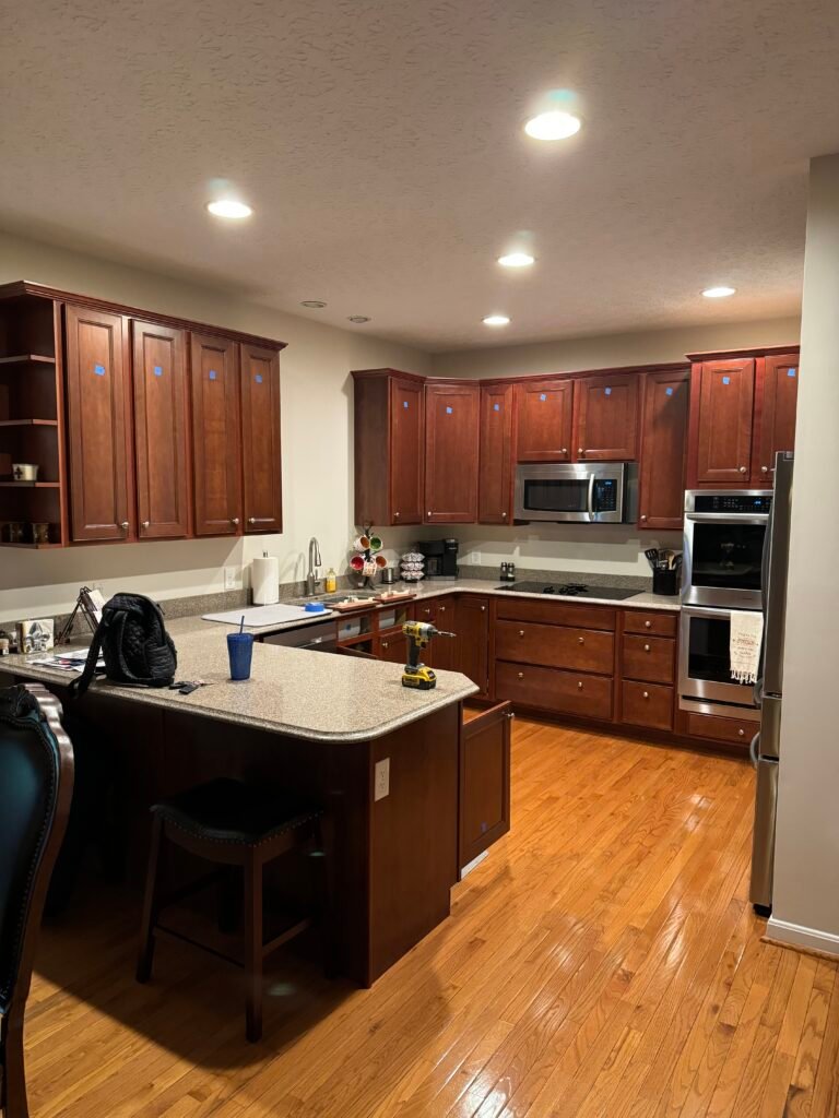 Modern kitchen with wooden cabinets, island, stainless steel appliances, and hardwood floors under bright ceiling lights.