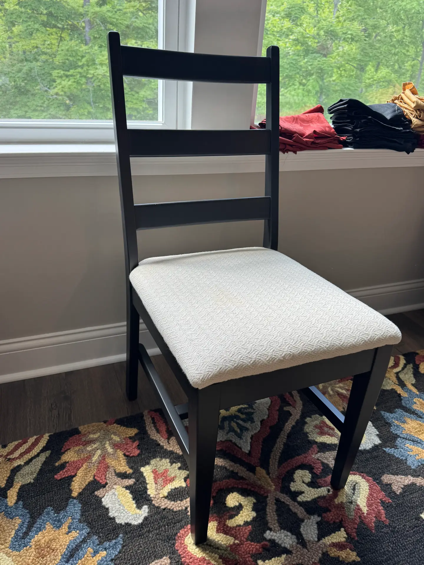 A black wooden chair with a white cushioned seat sits on a patterned rug near a window with a view of greenery. - My Personal Painter A black wooden chair with a white cushioned seat sits on a patterned rug near a window with a view of greenery.