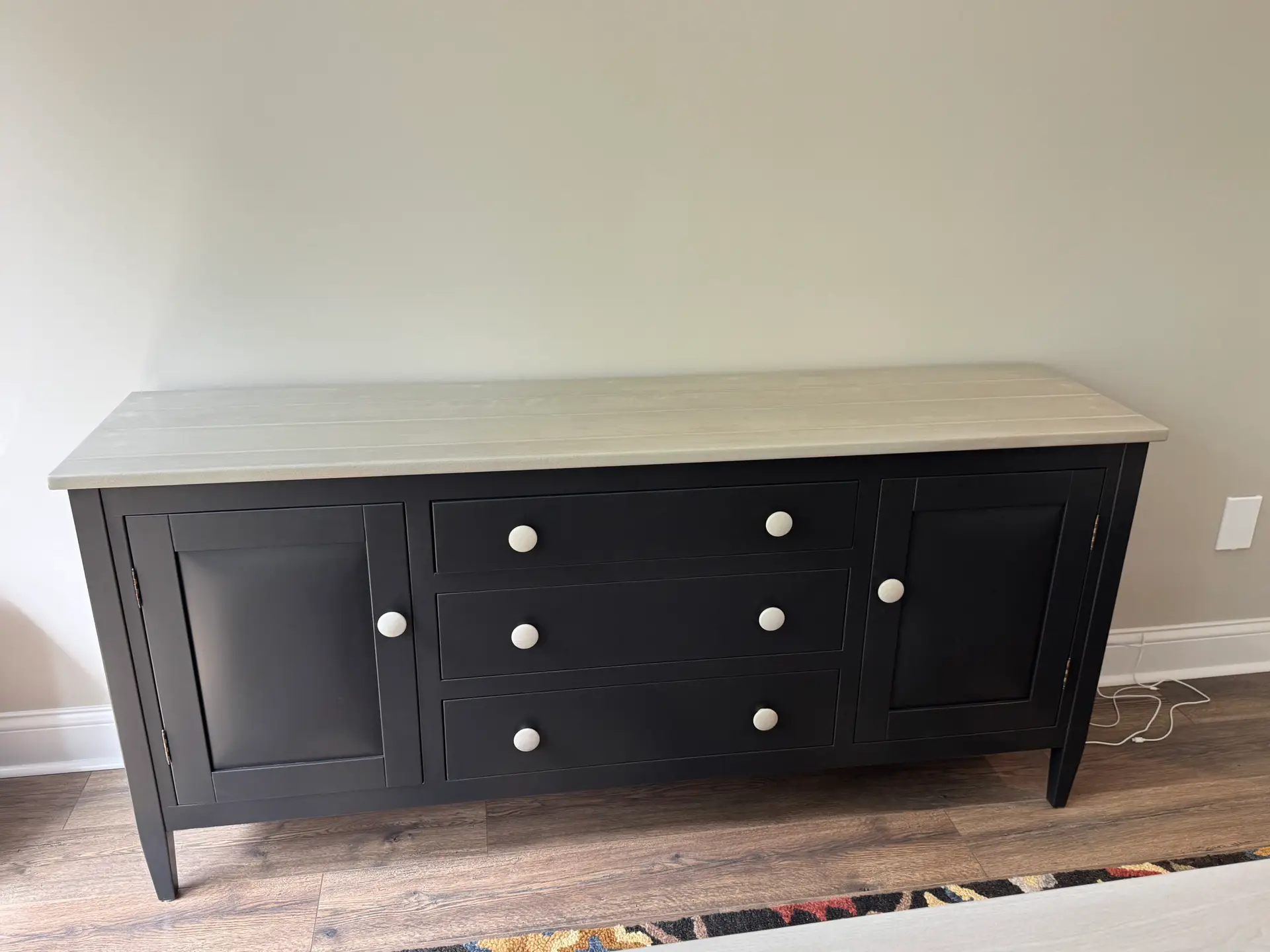 Black sideboard with white knobs, two doors, three drawers, against a light wall, on wood flooring.