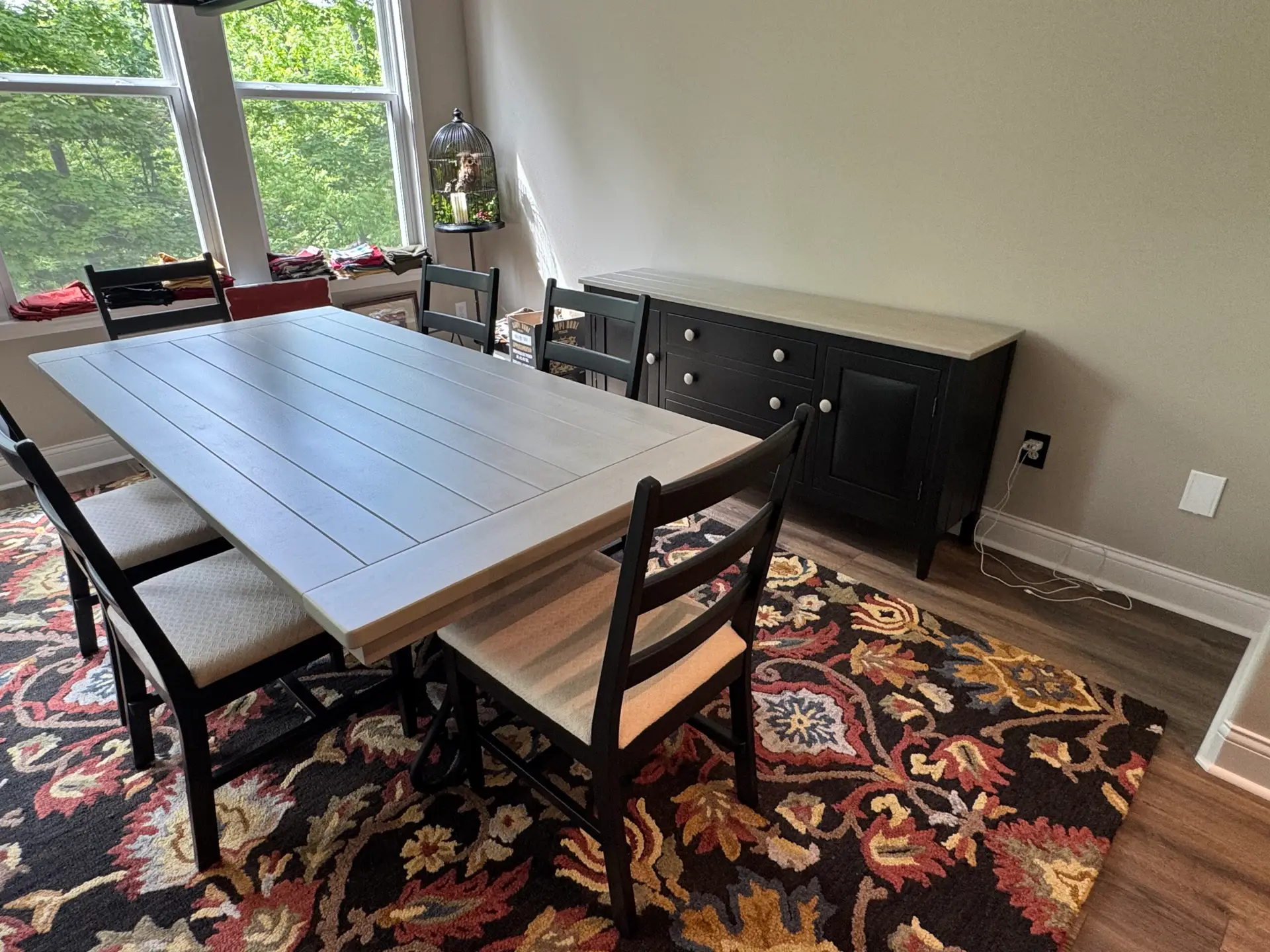 Dining room with a rectangular table, six chairs, a sideboard, floral rug, and large windows with greenery outside.
