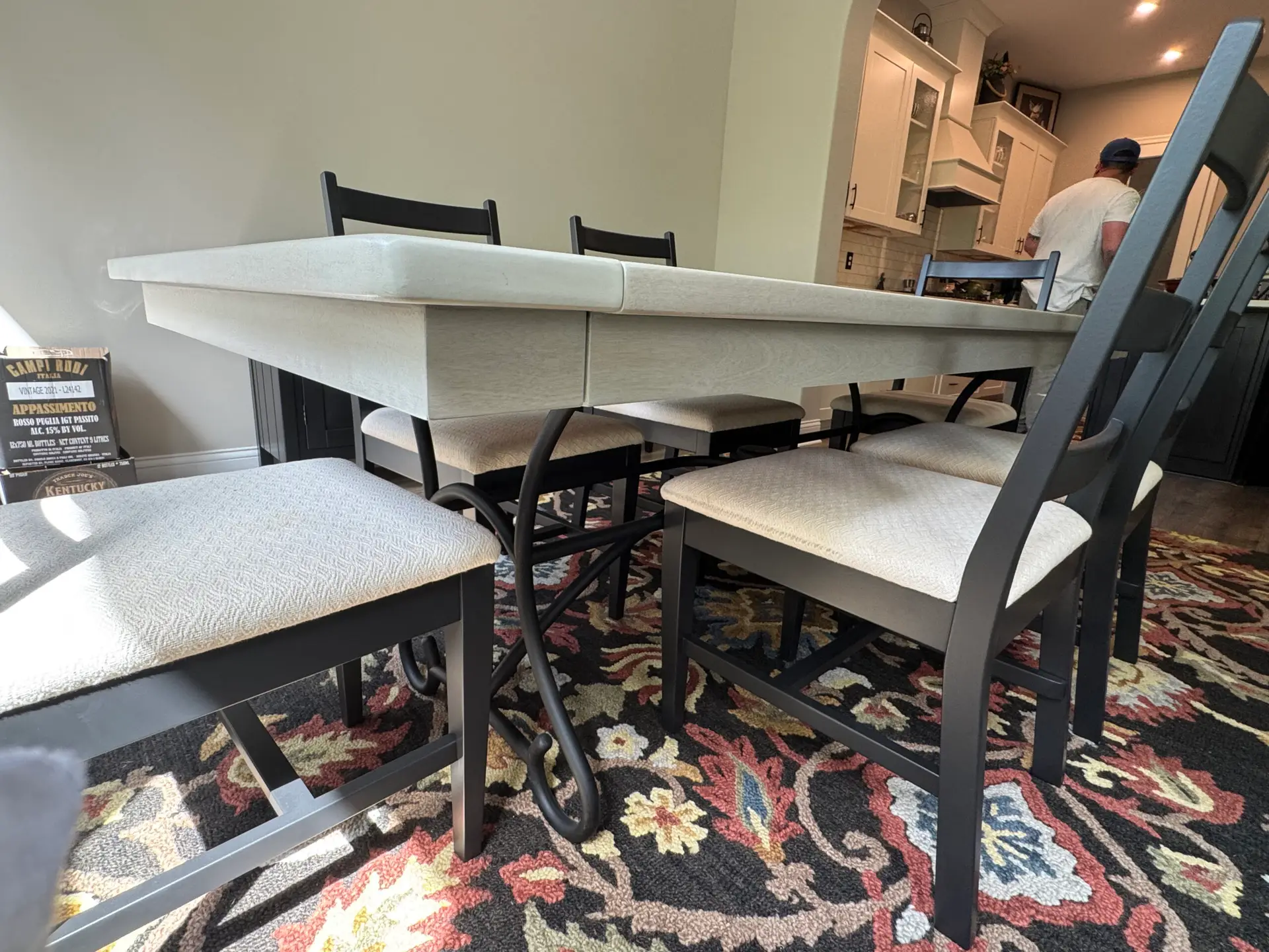 Dining table with six cushioned chairs on a floral rug; a person stands in the kitchen background. - My Personal Painter Dining table with six cushioned chairs on a floral rug; a person stands in the kitchen background.