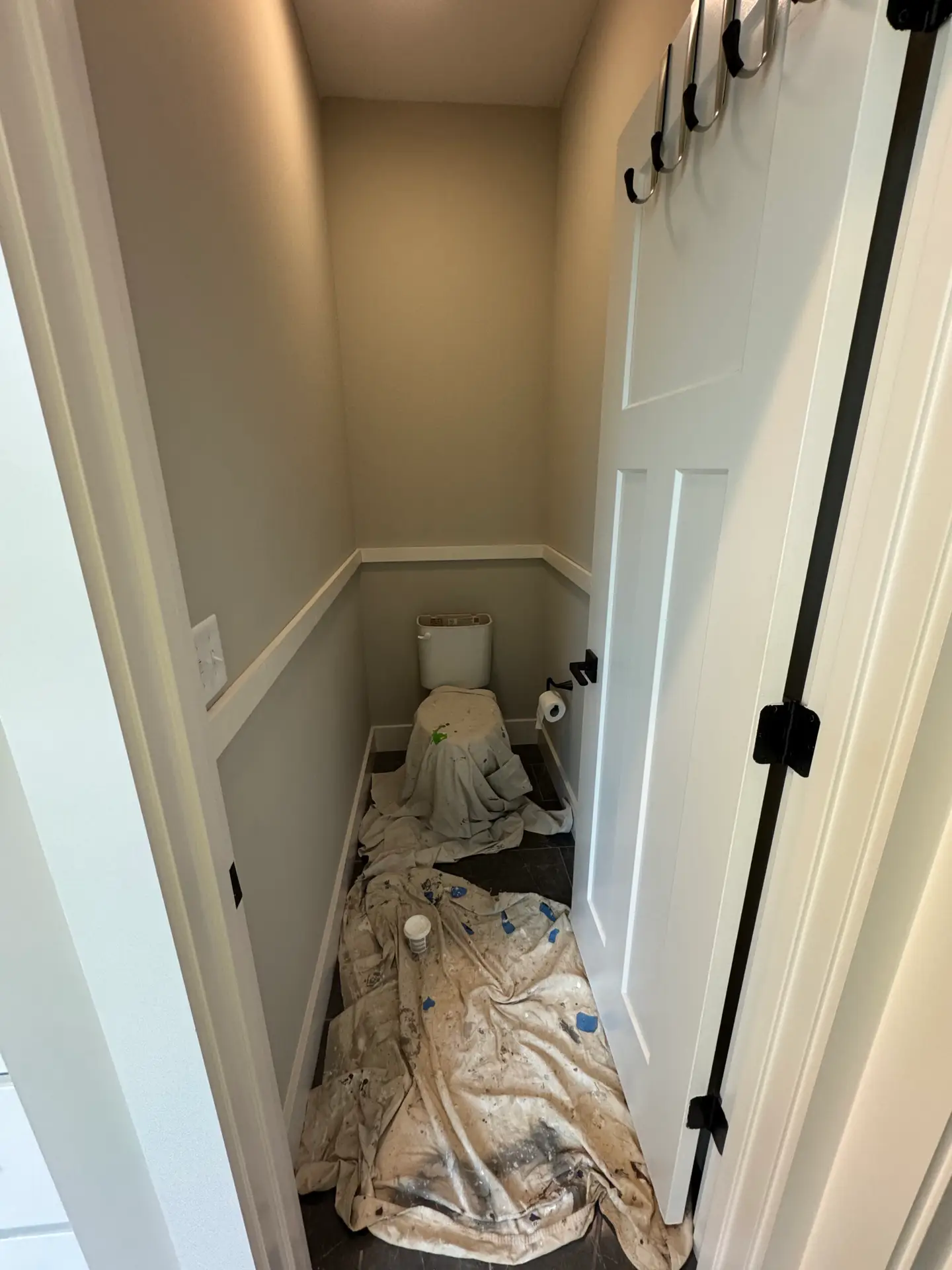 Small bathroom with a toilet, door open, tarps and paint supplies covering the floor, suggesting painting in progress.