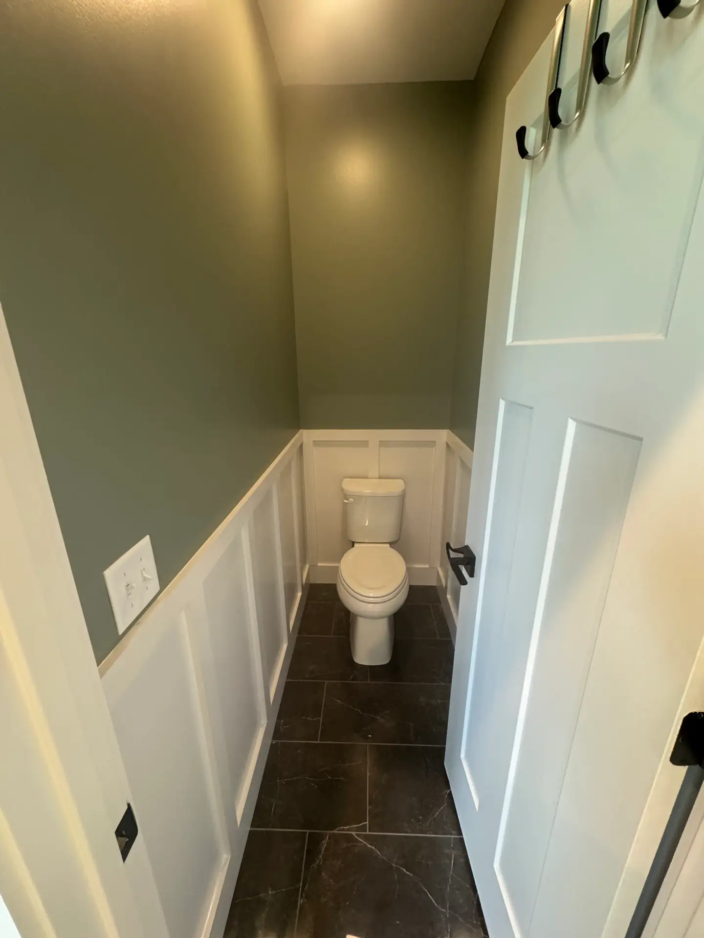Small bathroom with dark tile floor, green walls, white wainscoting, and a toilet centered at the far end.