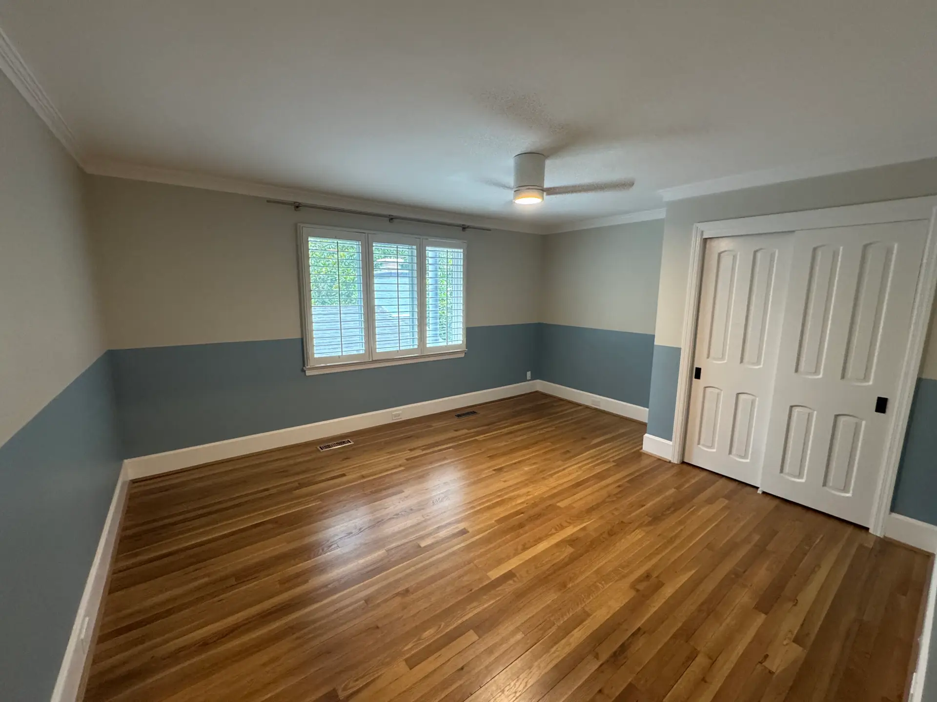 Empty room with hardwood floors, blue and gray walls, a window with blinds, ceiling fan, and white double closet doors.