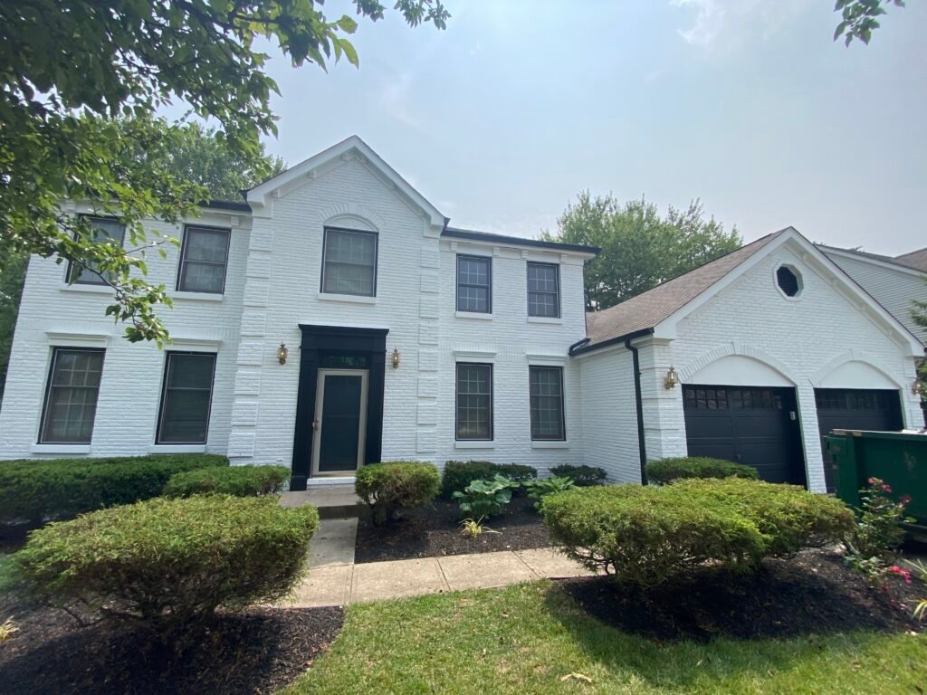 Two-story white brick house with black trim, black front door, and attached garage, surrounded by green shrubs and trees.