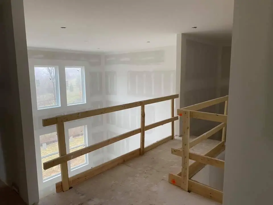 Unfinished loft area with wooden safety rails, drywall, and two large windows letting in natural light.