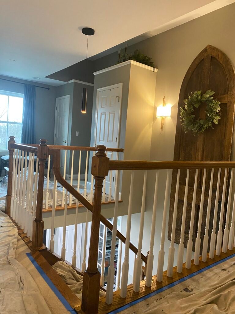 A hallway with wooden railings, two white doors, a wall sconce, and a wreath on a large rustic wooden wall piece.