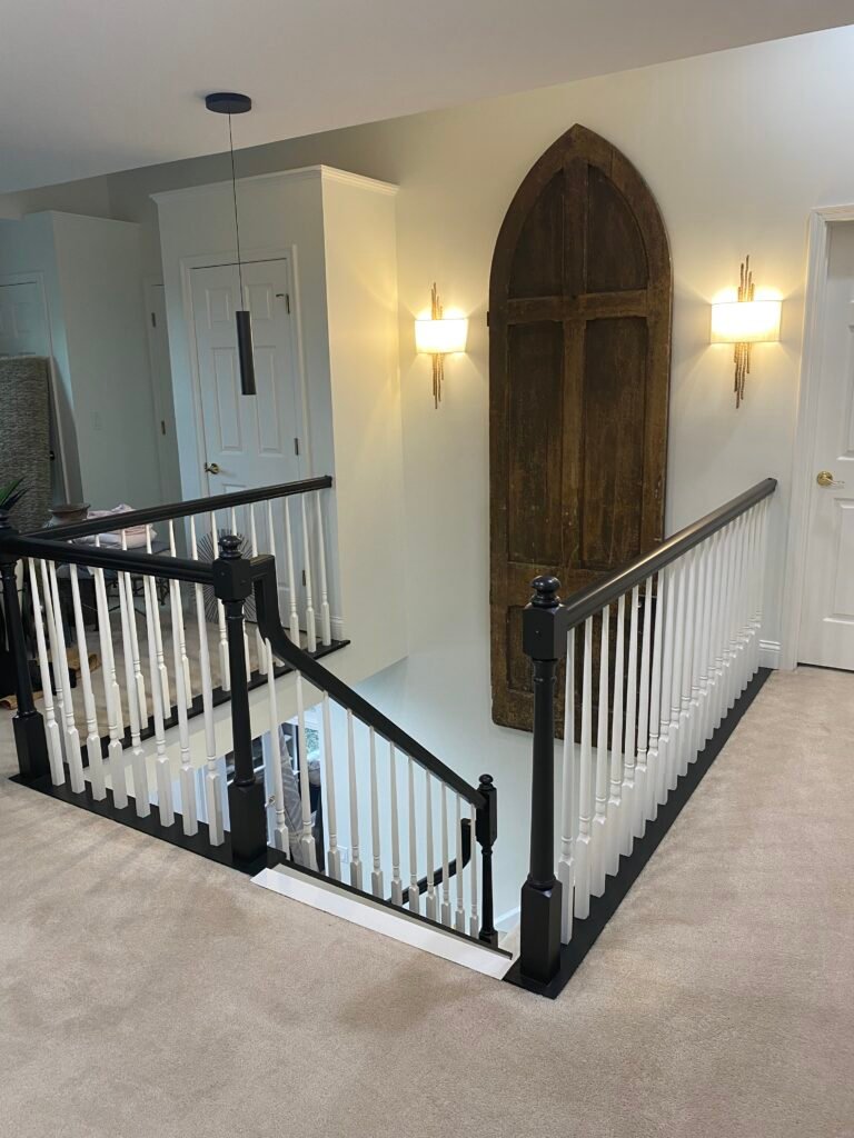 Wooden stairs with black-and-white railing, beige carpet, wall sconces, and a large arched wooden door on the wall.