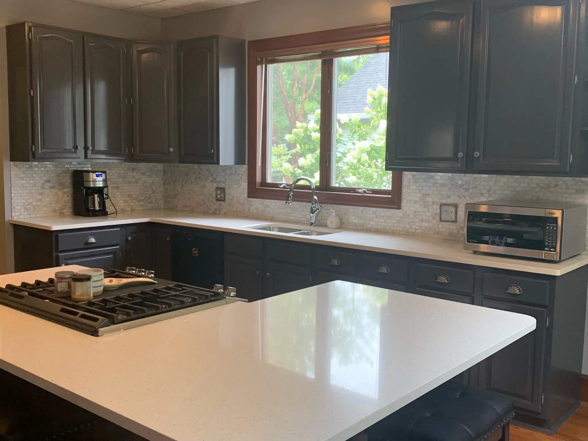 Modern kitchen with dark cabinets, white countertops, a large window, and appliances like a coffee maker and toaster oven. - My Personal Painter - My Personal Painter Kitchen with newly refinished charcoal-painted cabinets and white countertops