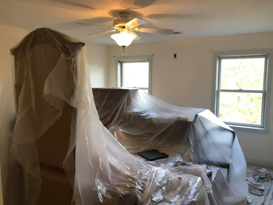 Furniture covered in plastic sheeting in a room with two windows and a ceiling fan. - My Personal Painter - My Personal Painter In progress: bathroom and bedroom painted light green