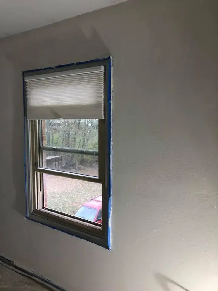 A window with a white pleated shade and blue painters tape around the frame on a plain wall. - My Personal Painter - My Personal Painter After: bedroom walls repaired and painted