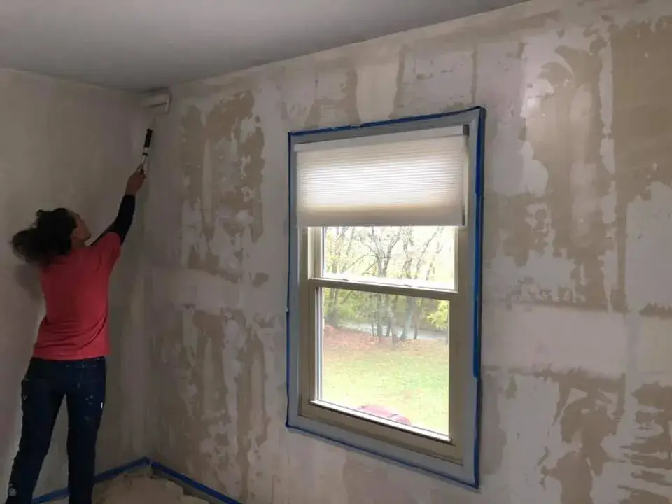 A person in a red shirt uses a paint roller to apply primer to a patched wall next to a window. - My Personal Painter - My Personal Painter In progress: bedroom walls repaired and painted