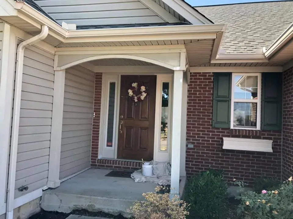 Front entrance of a brick and siding house with a brown door, floral wreath, and a paint bucket on the porch. - My Personal Painter - My Personal Painter In progress: brick and siding ranch: entry, doors and garage door