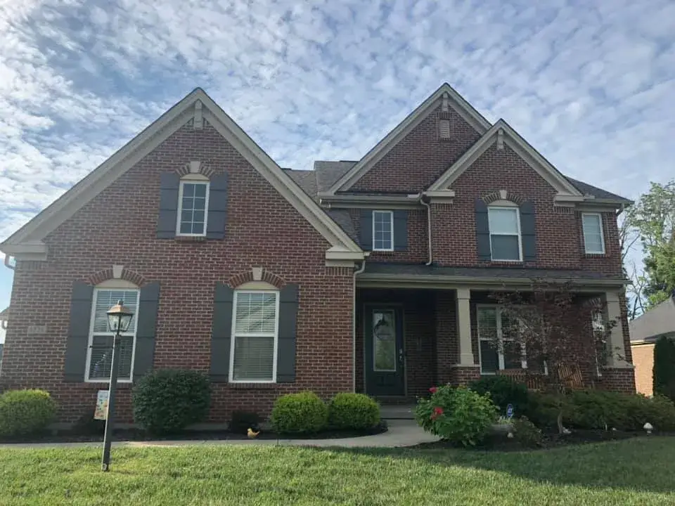 A two-story red brick house with dark shutters and a front porch under a partly cloudy sky. - My Personal Painter - My Personal Painter After: brick and tan siding house exterior painted