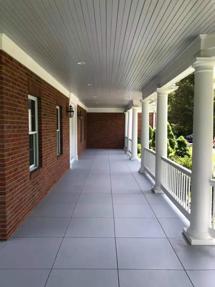 A covered porch with a red brick wall, grey tiled floor, and white columns. - My Personal Painter - My Personal Painter After: brick home: round wraparound porch and rear deck with brick columns