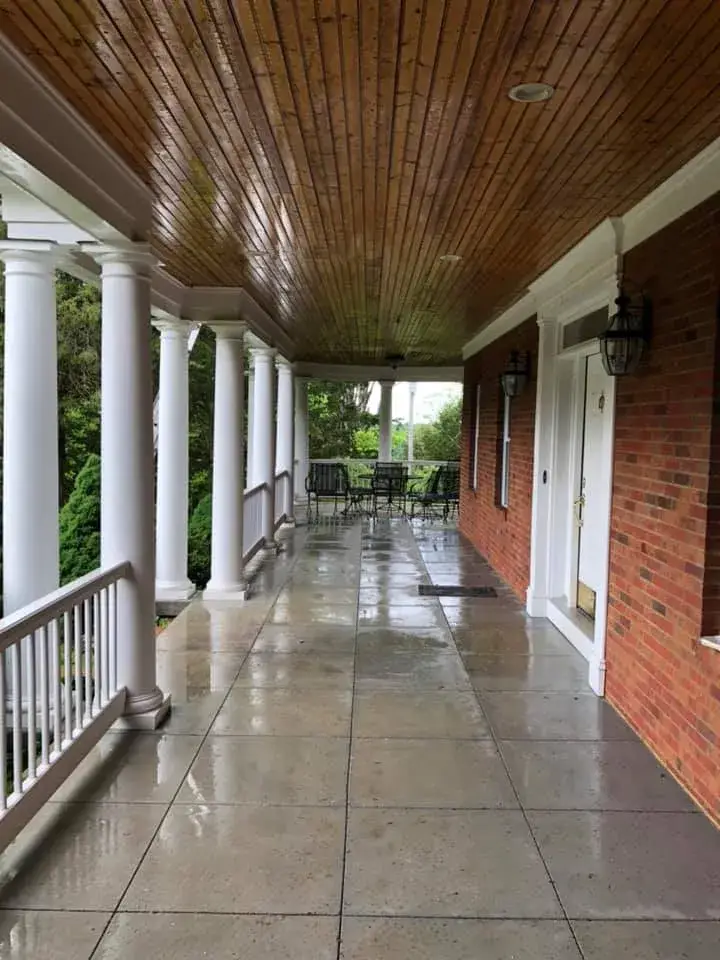 A covered porch with a tiled floor, white columns, and a wooden ceiling, overlooking a yard with patio furniture. - My Personal Painter - My Personal Painter Before: brick home: round wraparound porch and rear deck with brick columns