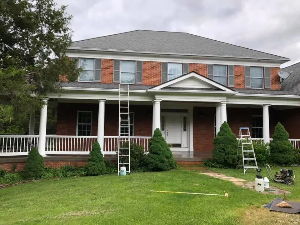 Two-story brick house with a covered porch, white columns, and ladders positioned for exterior maintenance. - My Personal Painter - My Personal Painter In progress: brick home: round wraparound porch and rear deck with brick columns