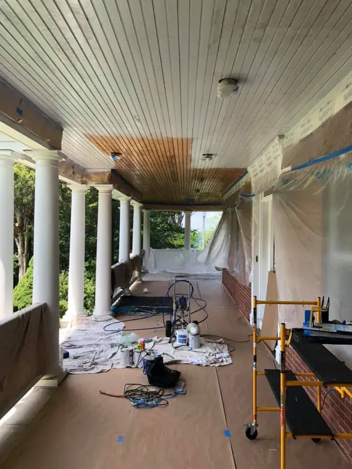 A porch undergoing renovation with protective plastic and paper covering the walls and floor. - My Personal Painter - My Personal Painter In progress: brick home: round wraparound porch and rear deck with brick columns