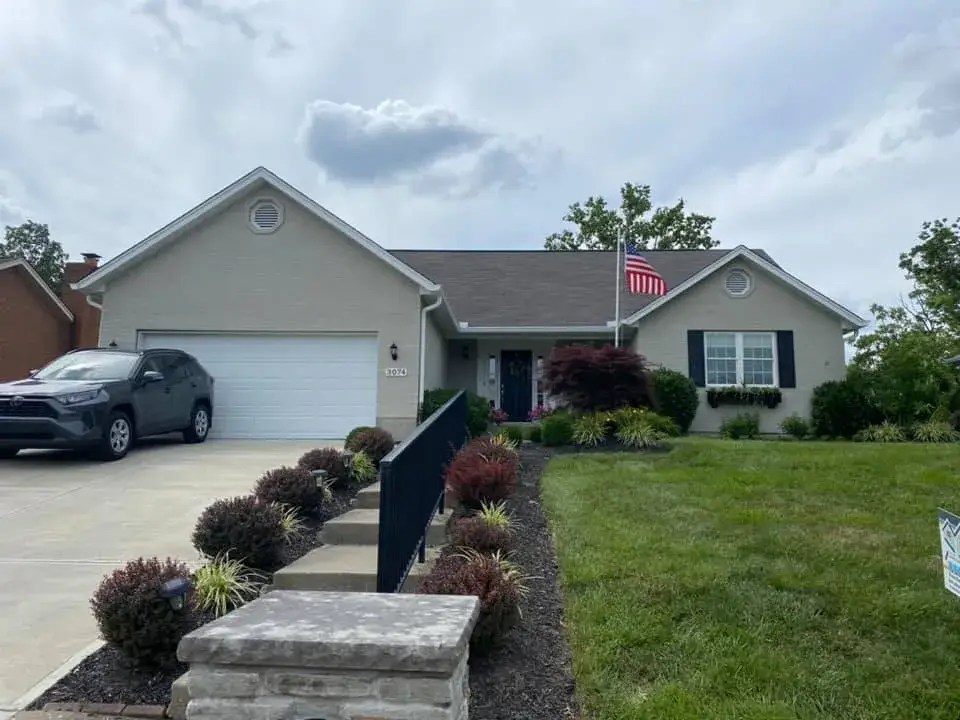 A beige single-story house with a white garage door, driveway, and American flag on the front lawn. - My Personal Painter - My Personal Painter After: brick ranch exterior painted light gray