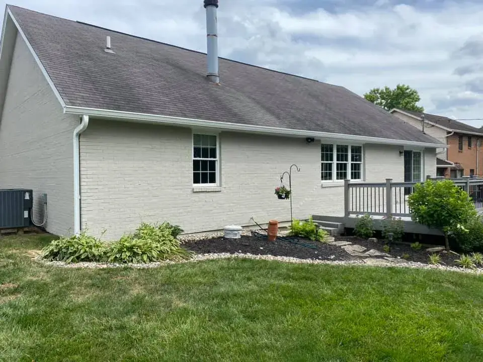 The back of a one-story brick house with a gray shingled roof, a chimney, and a small wooden deck. - My Personal Painter - My Personal Painter After: brick ranch exterior painted light gray