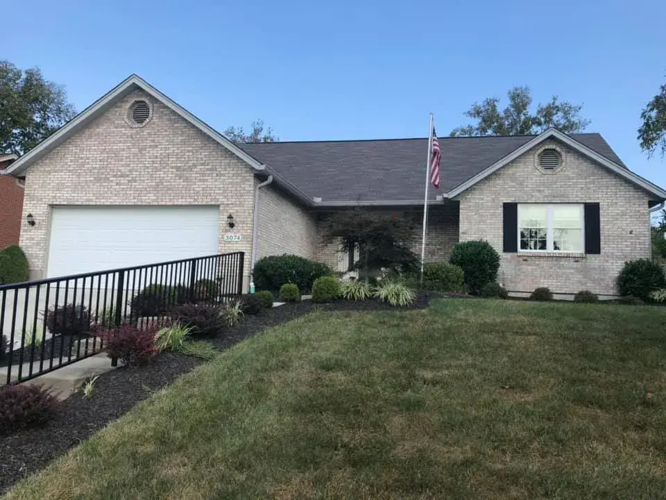 Single-story brick house with a two-car garage, a black railing along the driveway, and a front lawn. - My Personal Painter - My Personal Painter Before: brick ranch exterior painted light gray