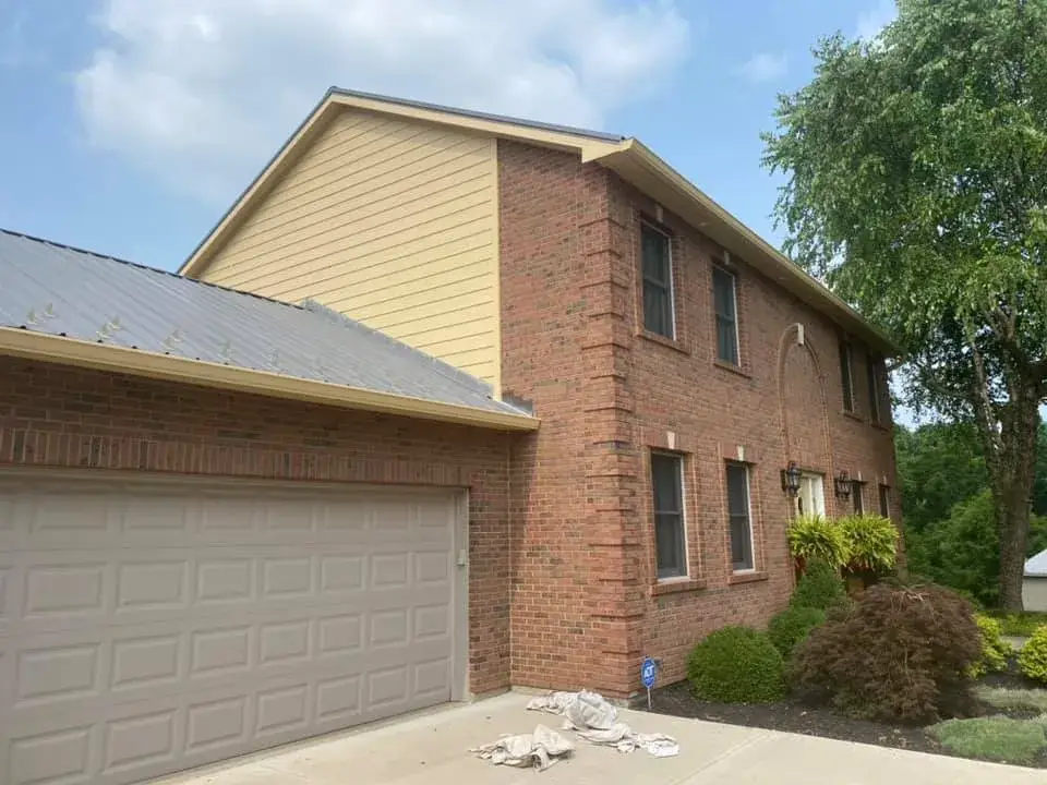 A two-story brick house with a metal roof over a tan garage door and tan siding on the upper gable. - My Personal Painter - My Personal Painter In progress: brick two-story house: siding and trim painted