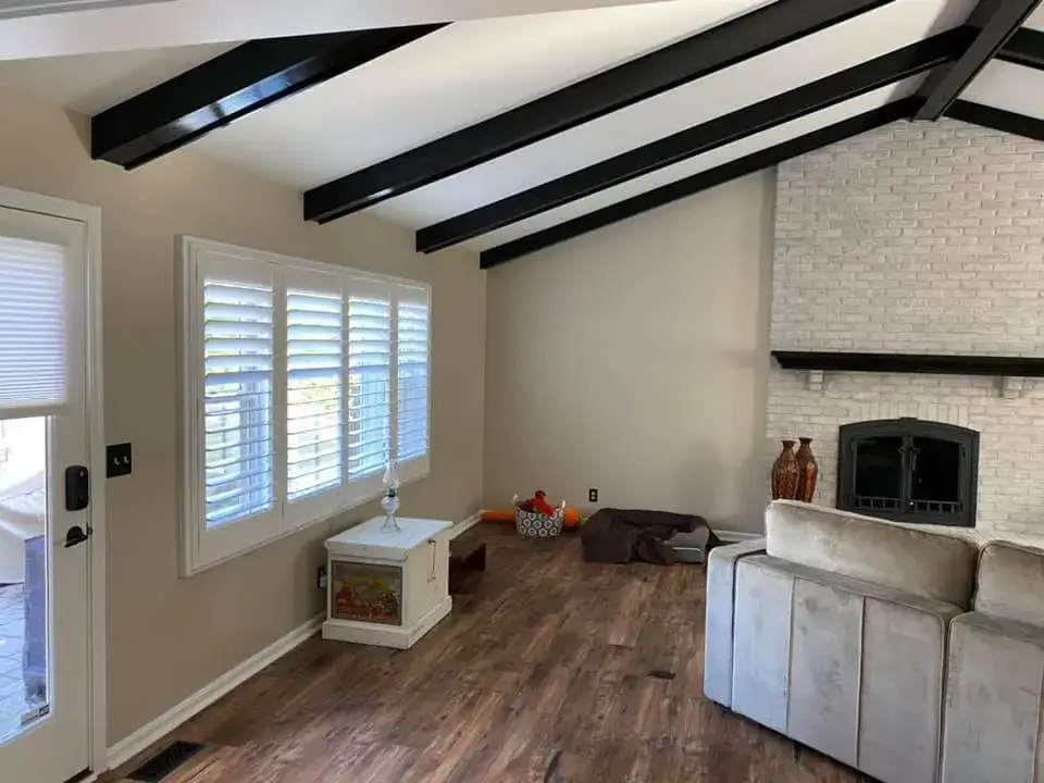 Living room with a brick fireplace, black ceiling beams, wood-look flooring, and white plantation shutters. - My Personal Painter - My Personal Painter After: ceiling beams painted black, brick fireplace whitewashed, other rooms