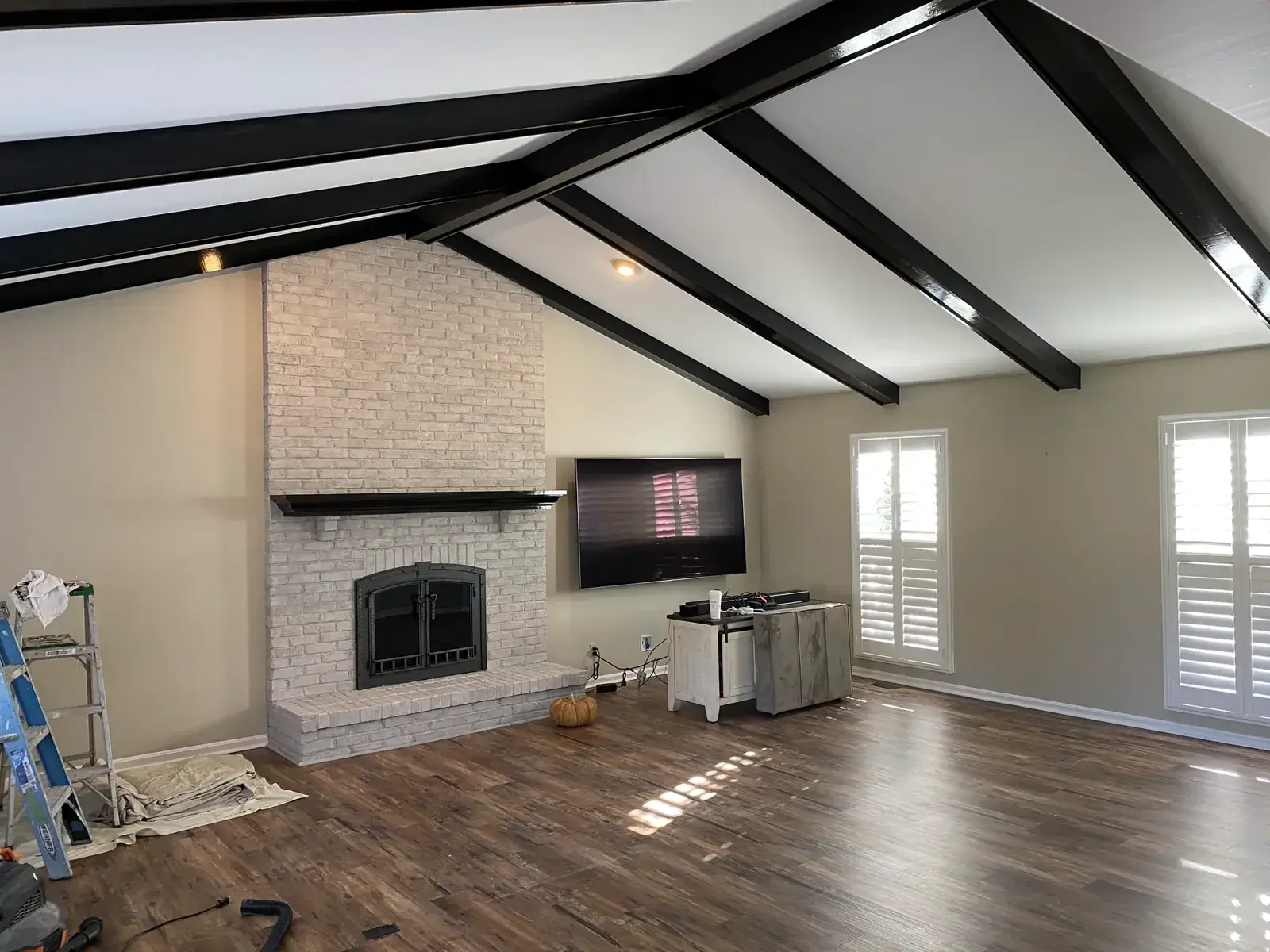 Living room with a white brick fireplace, black ceiling beams, wood-look flooring, and a mounted television. - My Personal Painter - My Personal Painter In progress: ceiling beams painted black, brick fireplace whitewashed, other rooms