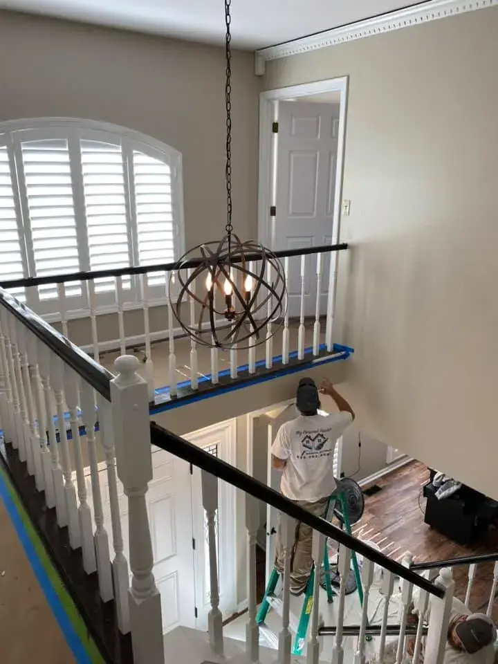 A painter on a ladder works on the wall of a two-story foyer near a large spherical chandelier. - My Personal Painter - My Personal Painter In progress: ceiling beams painted black, brick fireplace whitewashed, other rooms