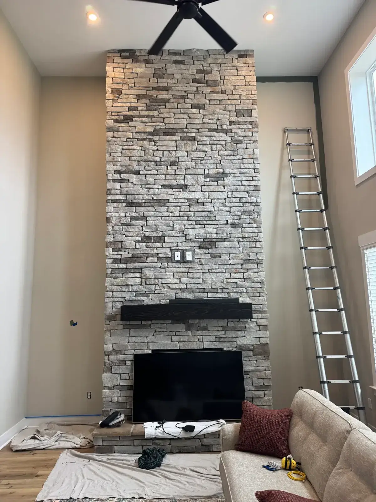 Floor-to-ceiling stone fireplace with a black mantel and mounted television, next to an extended ladder. - My Personal Painter - My Personal Painter In progress: green walls beside a tall stone fireplace, brown and copper bedroom accent wall