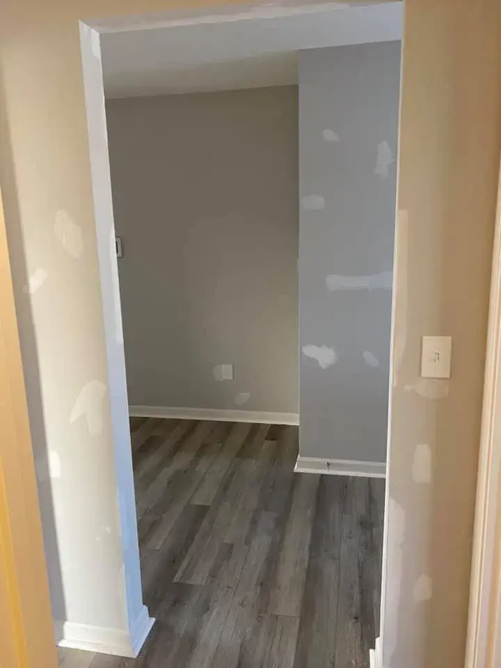 A doorway leading to a gray room with wood-look flooring and patches of white spackle on the walls. - My Personal Painter - My Personal Painter In progress: interior walls patched and painted gray