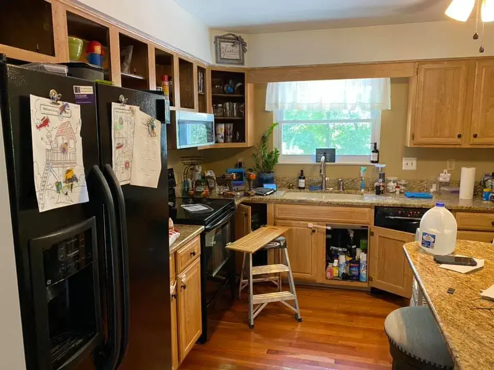 Kitchen with a black refrigerator, wooden cabinets, a step stool, and an open cabinet door under the sink. - My Personal Painter - My Personal Painter Before: kitchen cabinets painted white, granite counters