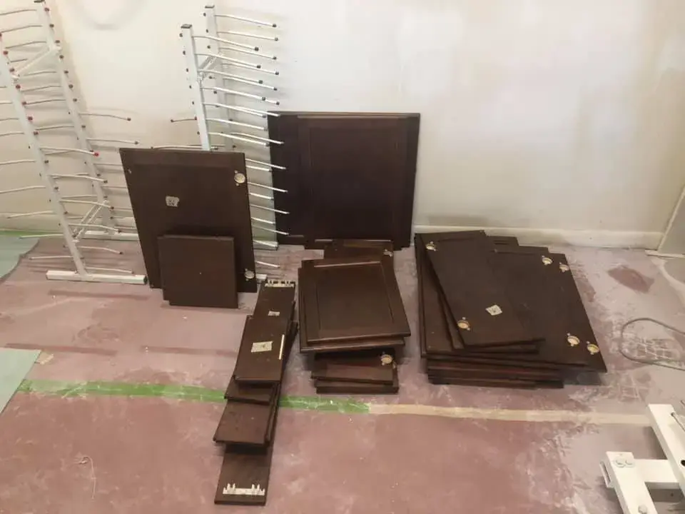 Stacks of dark wood cabinet doors and drawer fronts on a floor near two metal drying racks. - My Personal Painter - My Personal Painter In progress: kitchen cherry cabinets painted black