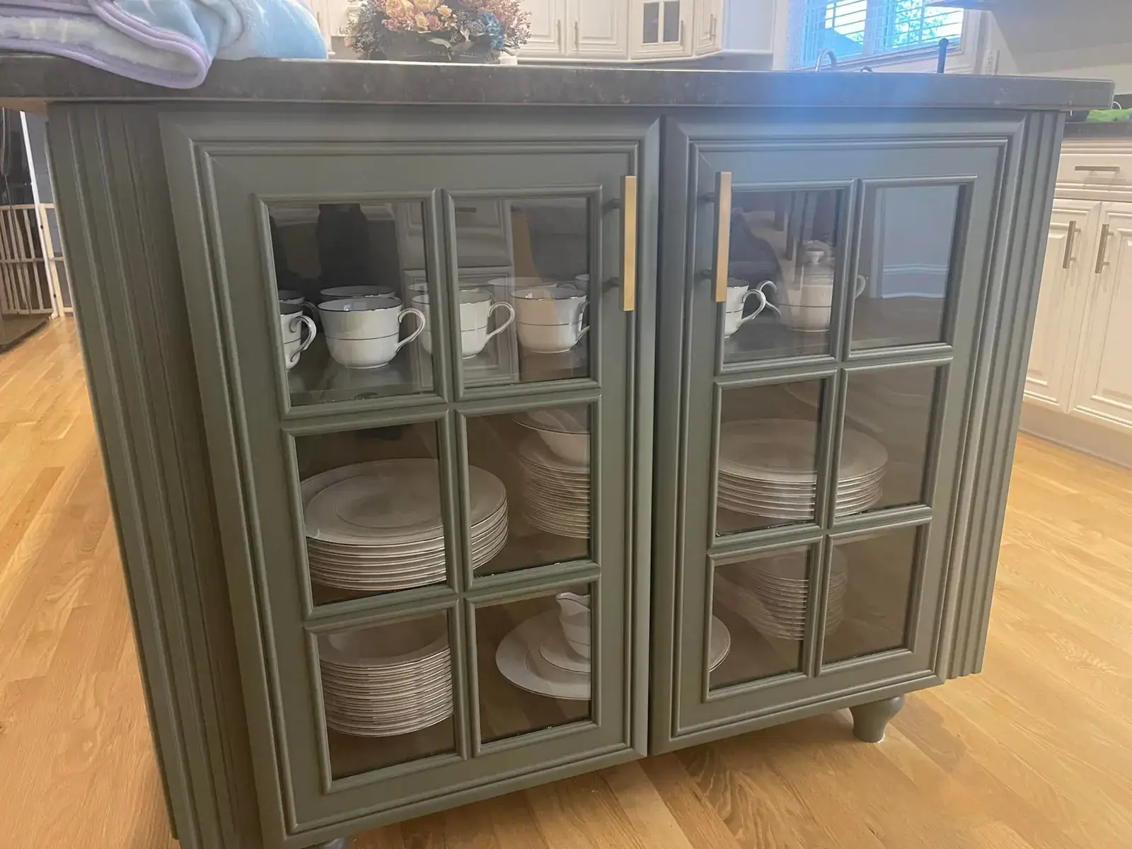 Sage green kitchen island with glass-front cabinets displaying stacks of white plates and teacups. - My Personal Painter - My Personal Painter After: kitchen cherry cabinets to white with a green island, two-story great room, dining room and foyer