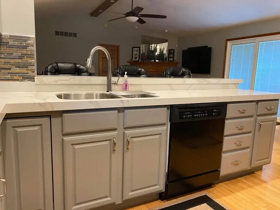 Kitchen island with a white countertop, stainless steel sink, and black dishwasher in front of grey cabinets. - My Personal Painter - My Personal Painter After: kitchen oak cabinets painted gray, vaulted ceiling with beam