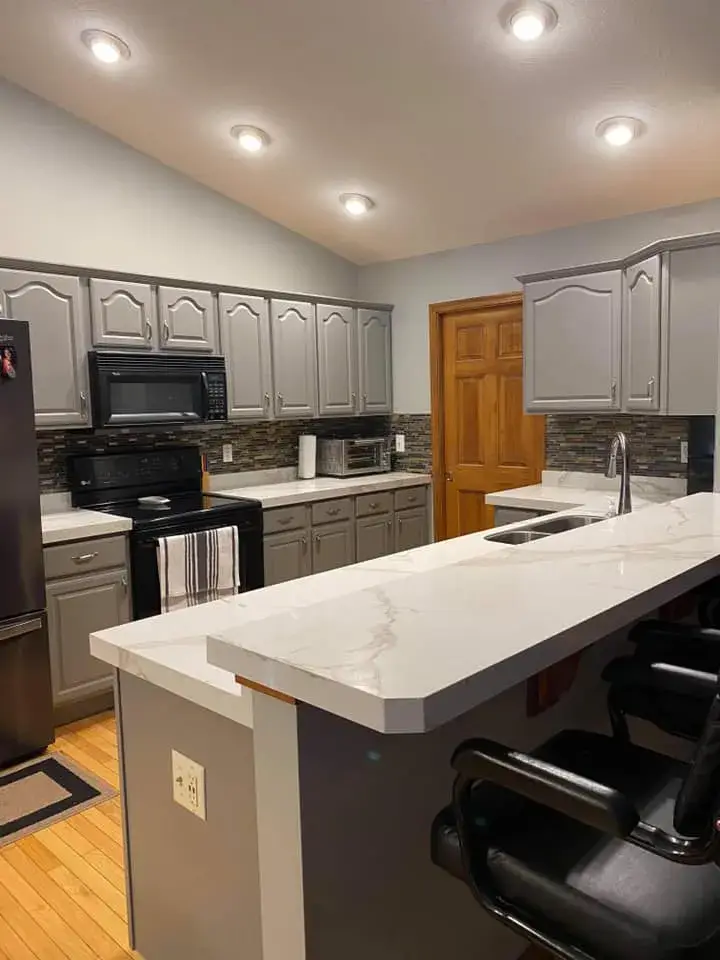 A kitchen with grey cabinets, a marble-look countertop, a black stove, and two black bar stools. - My Personal Painter - My Personal Painter After: kitchen oak cabinets painted gray, vaulted ceiling with beam