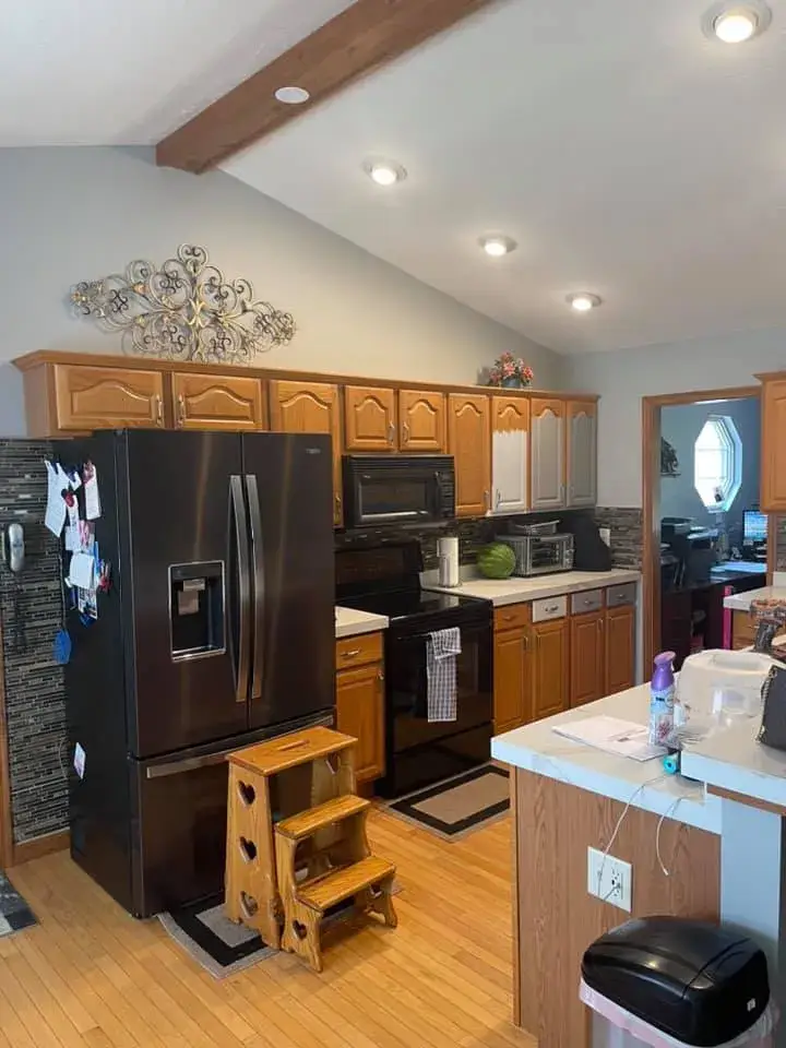 Kitchen with oak cabinets, a black refrigerator, a black stove, and a wooden step stool in the foreground. - My Personal Painter - My Personal Painter Before: kitchen oak cabinets painted gray, vaulted ceiling with beam