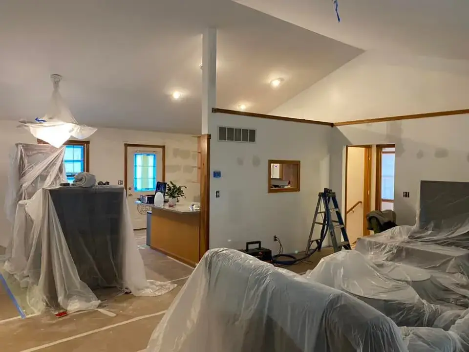 A room undergoing renovations, with furniture and fixtures covered in protective plastic sheeting. - My Personal Painter - My Personal Painter In progress: kitchen oak cabinets to white uppers and blue-gray lowers