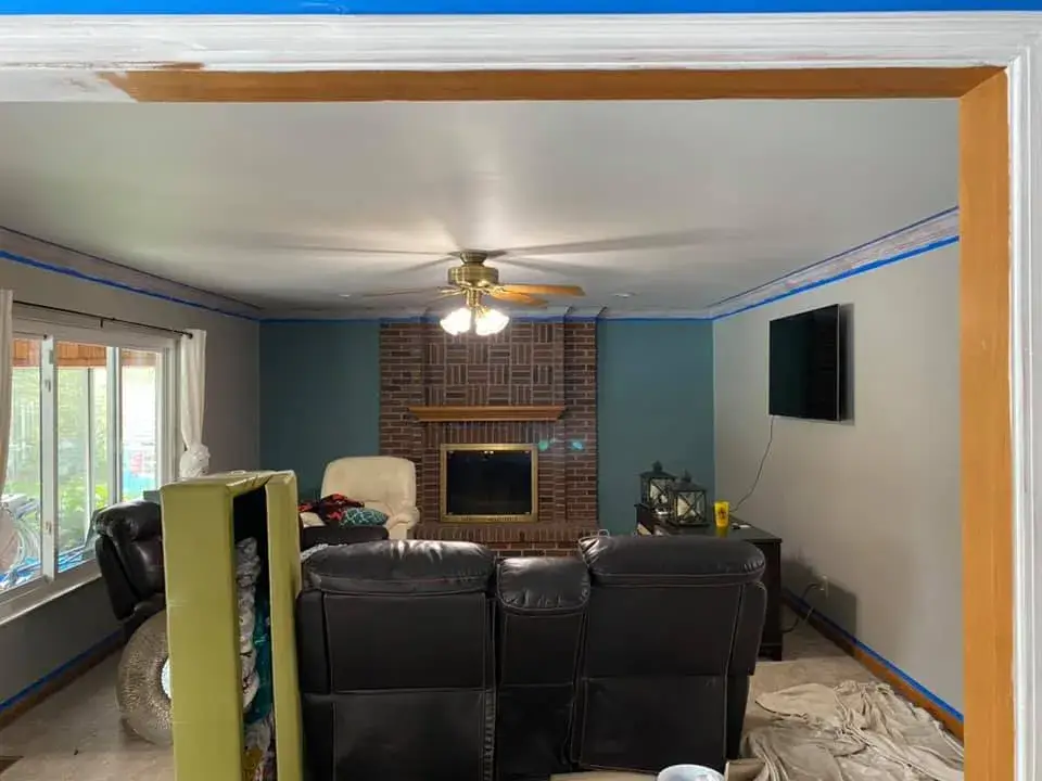 Living room featuring a brick fireplace, leather furniture, and blue painters tape along the ceiling and trim. - My Personal Painter - My Personal Painter Before: kitchen painted white, living room and dining room repainted
