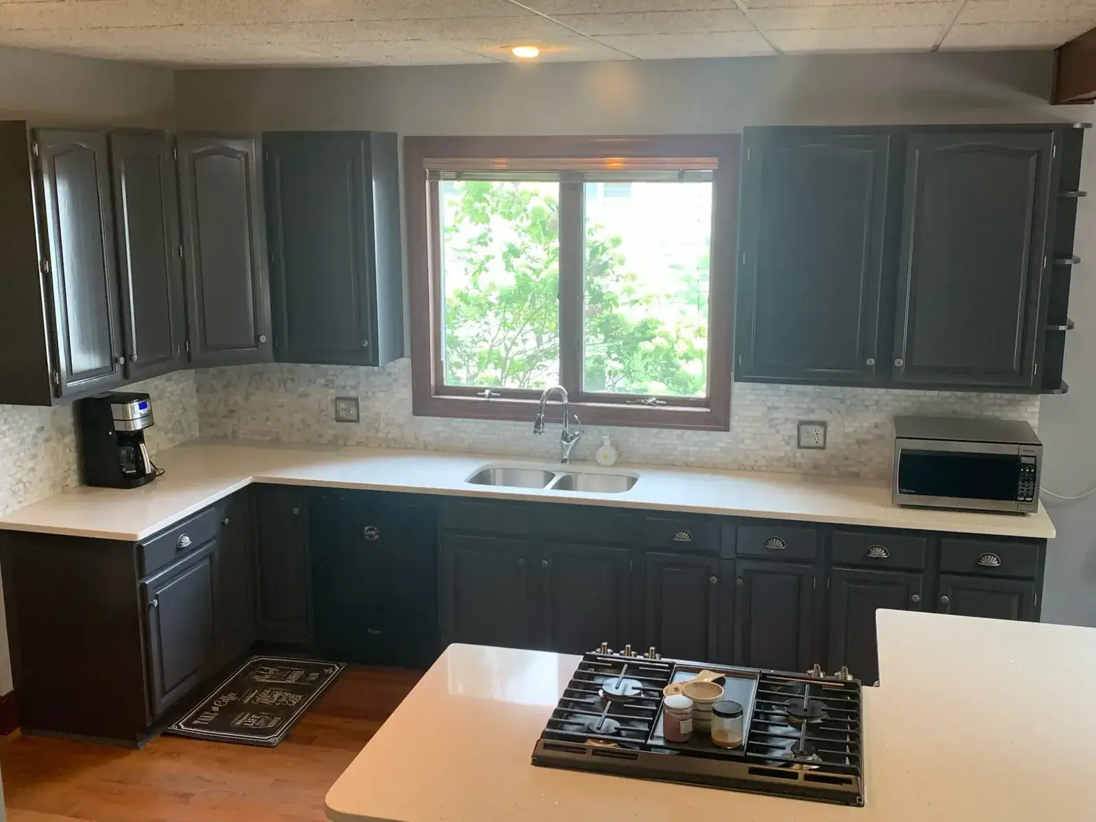 Kitchen with dark cabinets, white countertops, a double sink under a window, and a gas cooktop on the island. - My Personal Painter - My Personal Painter After: kitchen wood cabinets painted dark