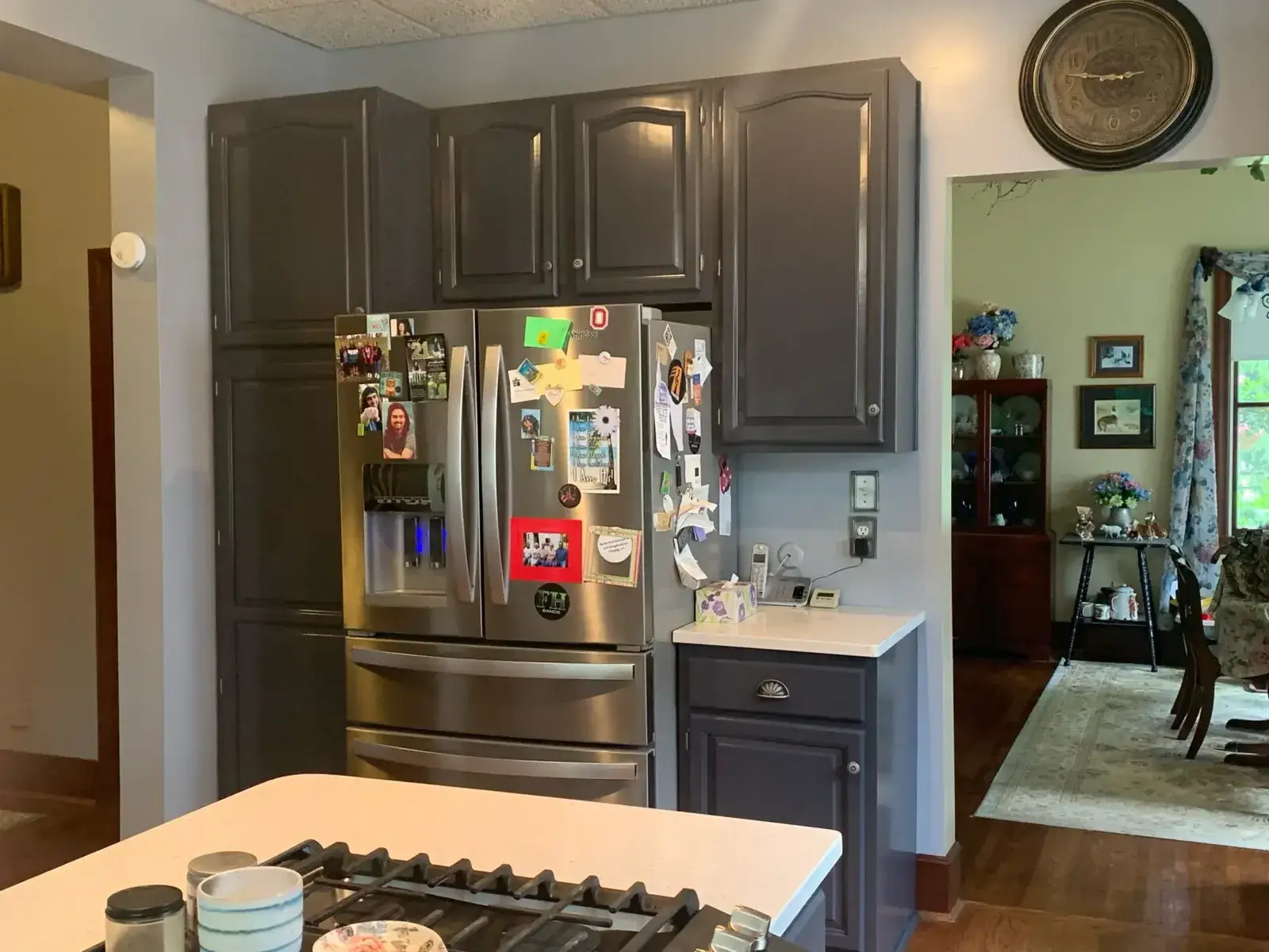 A stainless steel refrigerator covered in magnets and photos sits between dark gray kitchen cabinets. - My Personal Painter - My Personal Painter After: kitchen wood cabinets painted dark