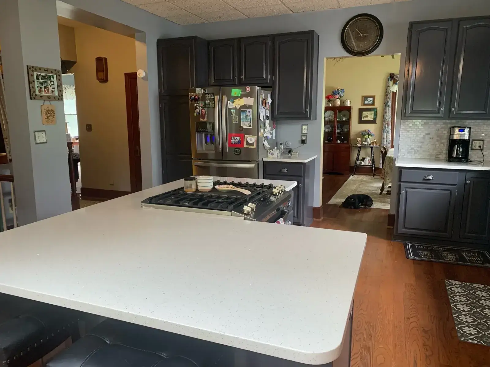 A kitchen with a large island, a gas cooktop, dark cabinets, and a stainless steel refrigerator. - My Personal Painter - My Personal Painter After: kitchen wood cabinets painted dark