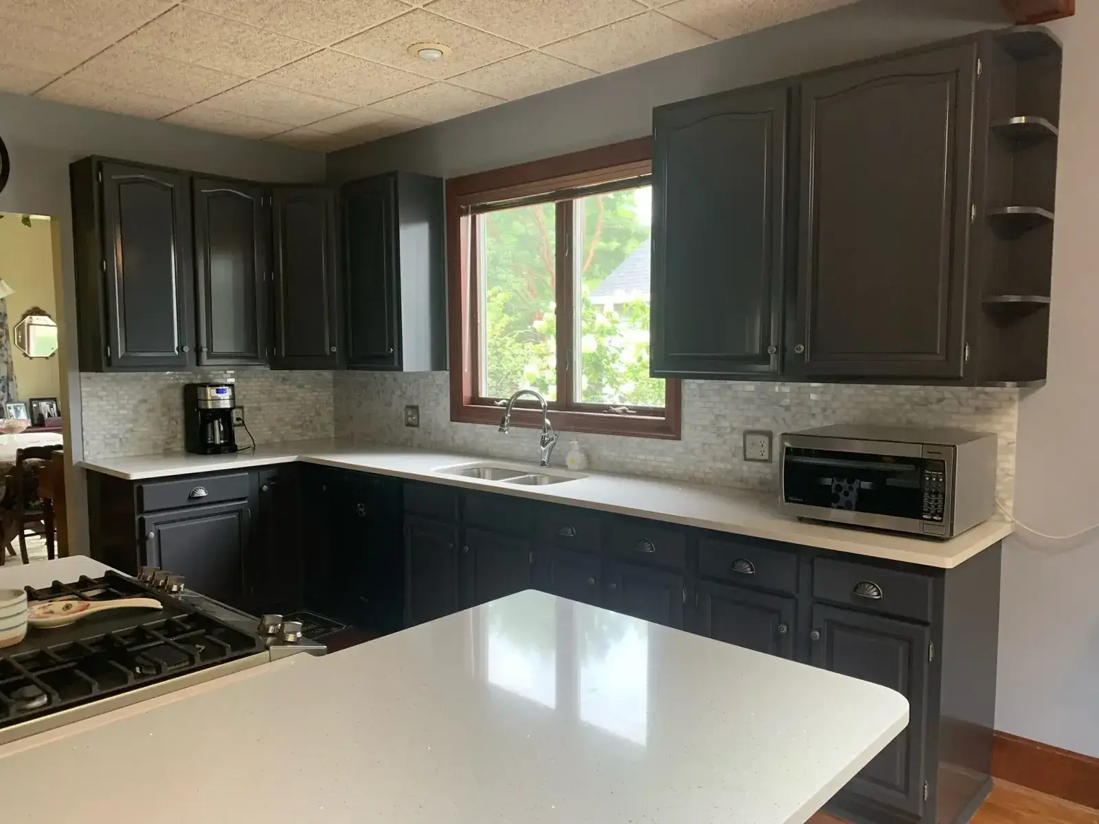 Kitchen with dark gray cabinets, white countertops, a central island, and a window above the sink. - My Personal Painter - My Personal Painter After: kitchen wood cabinets painted dark