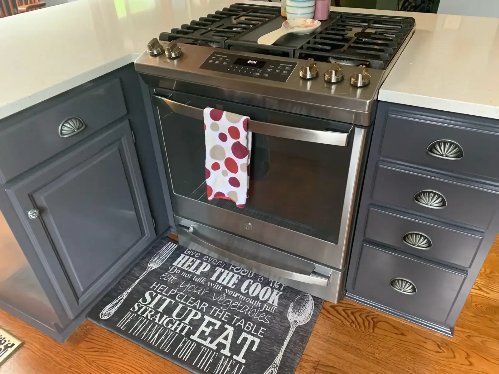 Stainless steel gas range with a polka-dot towel hanging on the oven door and a kitchen mat with dining-related text. - My Personal Painter - My Personal Painter After: kitchen wood cabinets painted dark