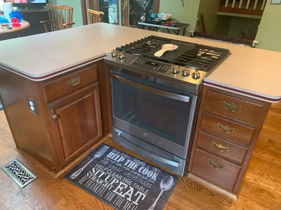 Kitchen island featuring a stainless steel stove with an oven and a floor mat with cooking-themed text. - My Personal Painter - My Personal Painter Before: kitchen wood cabinets painted dark