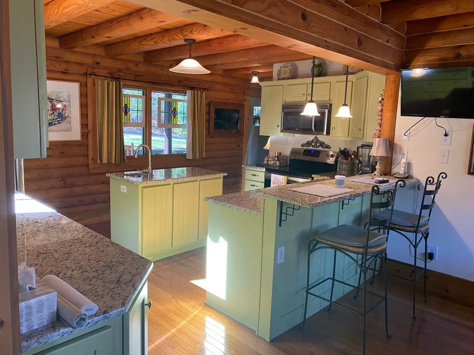 A rustic kitchen featuring log walls, light green cabinets, granite countertops, and a breakfast bar with two stools. - My Personal Painter - My Personal Painter After: log cabin kitchen painted green