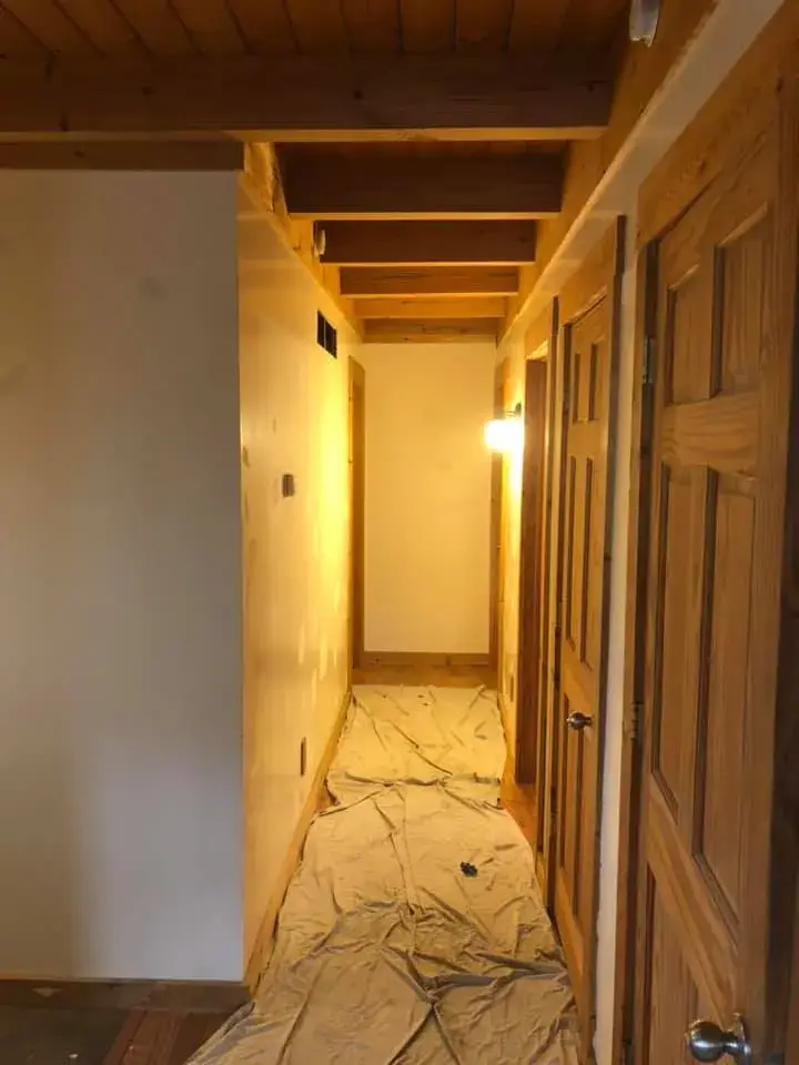 A narrow hallway with exposed wooden ceiling beams and a drop cloth covering the floor. - My Personal Painter - My Personal Painter In progress: log home with timber ceilings: walls painted