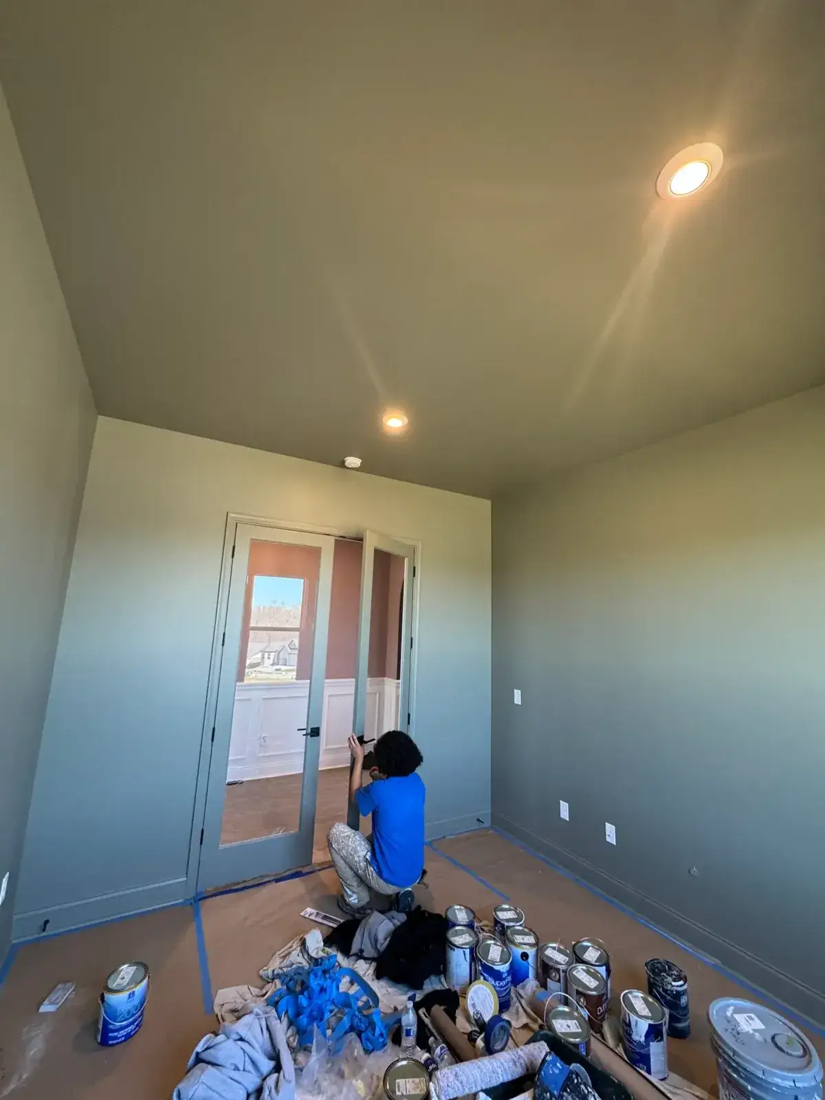 A person in a blue shirt kneels on a drop cloth in a sage green room, taking a photo of a glass-paneled door. - My Personal Painter - My Personal Painter In progress: new-build house: black archway, accent walls and doors